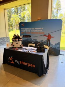 FullSizeRender | mysherpas.ai | Purpose Built Business Lending AI Platform MySherpas booth with a curved blue banner reading 'Pioneering the new era in lending technology. Meet your trusted guide.' Plush toys on a cooler and brochures on the table.